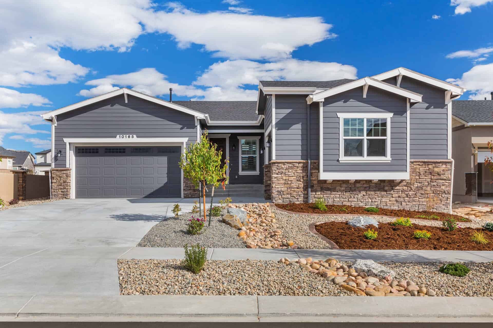 Single-story modern house with gray siding, stone accents, and a two-car garage; the front yard is beautifully landscaped by a professional landscaper with rocks and mulch under a blue sky.