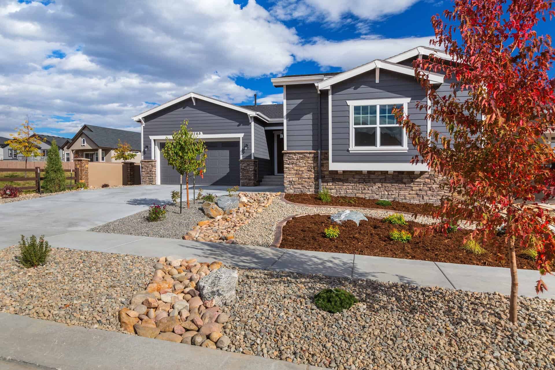 A modern single-story house with gray siding, stone accents, and a landscaped front yard designed by a professional landscaper, featuring rocks, mulch, and young trees under a partly cloudy sky.