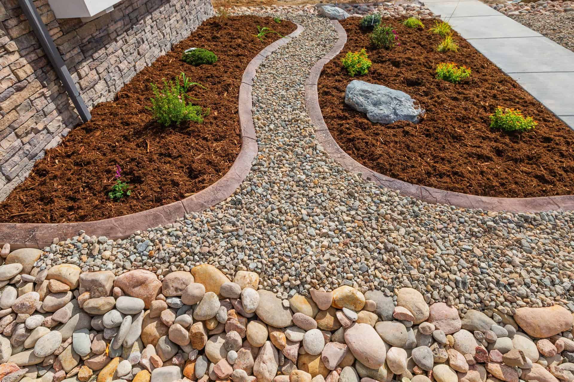 Curved gravel pathway, expertly crafted by a landscaper, is bordered by mulch, rocks, and small plants, running alongside a stone house exterior and a concrete sidewalk.
