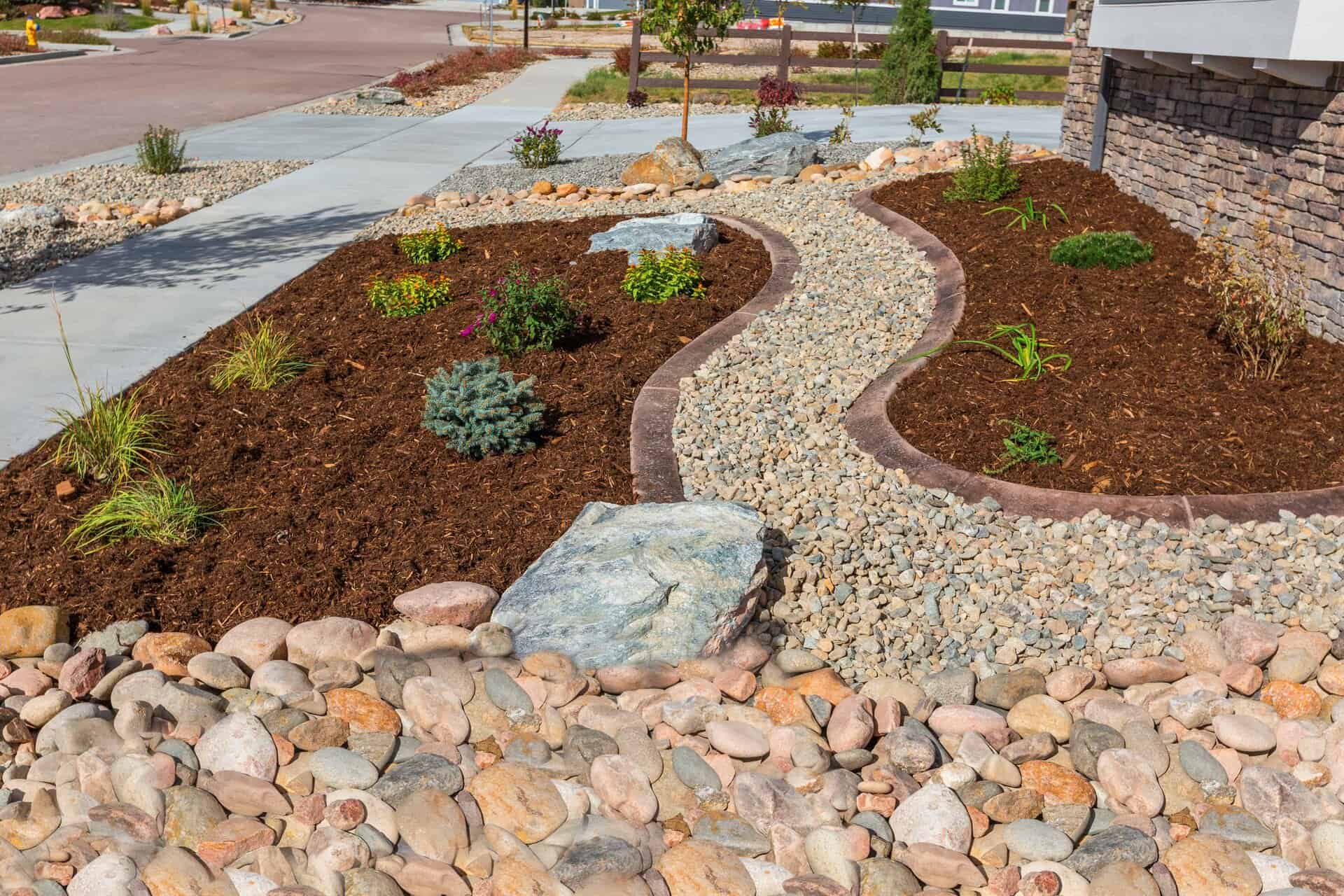 A landscaper-designed xeriscaped front yard featuring mulch, decorative rocks, small shrubs, and a stone border next to a sidewalk and street.