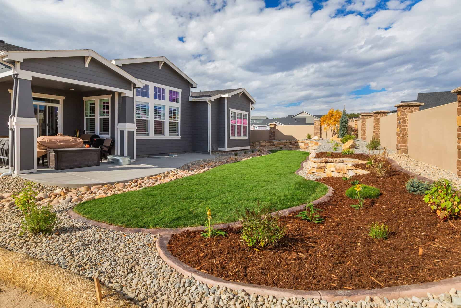 Single-story house with large windows, a covered patio, and a backyard expertly designed by a landscaper—featuring a lush lawn, mulch beds, stone edging, and gravel areas under a partly cloudy sky.