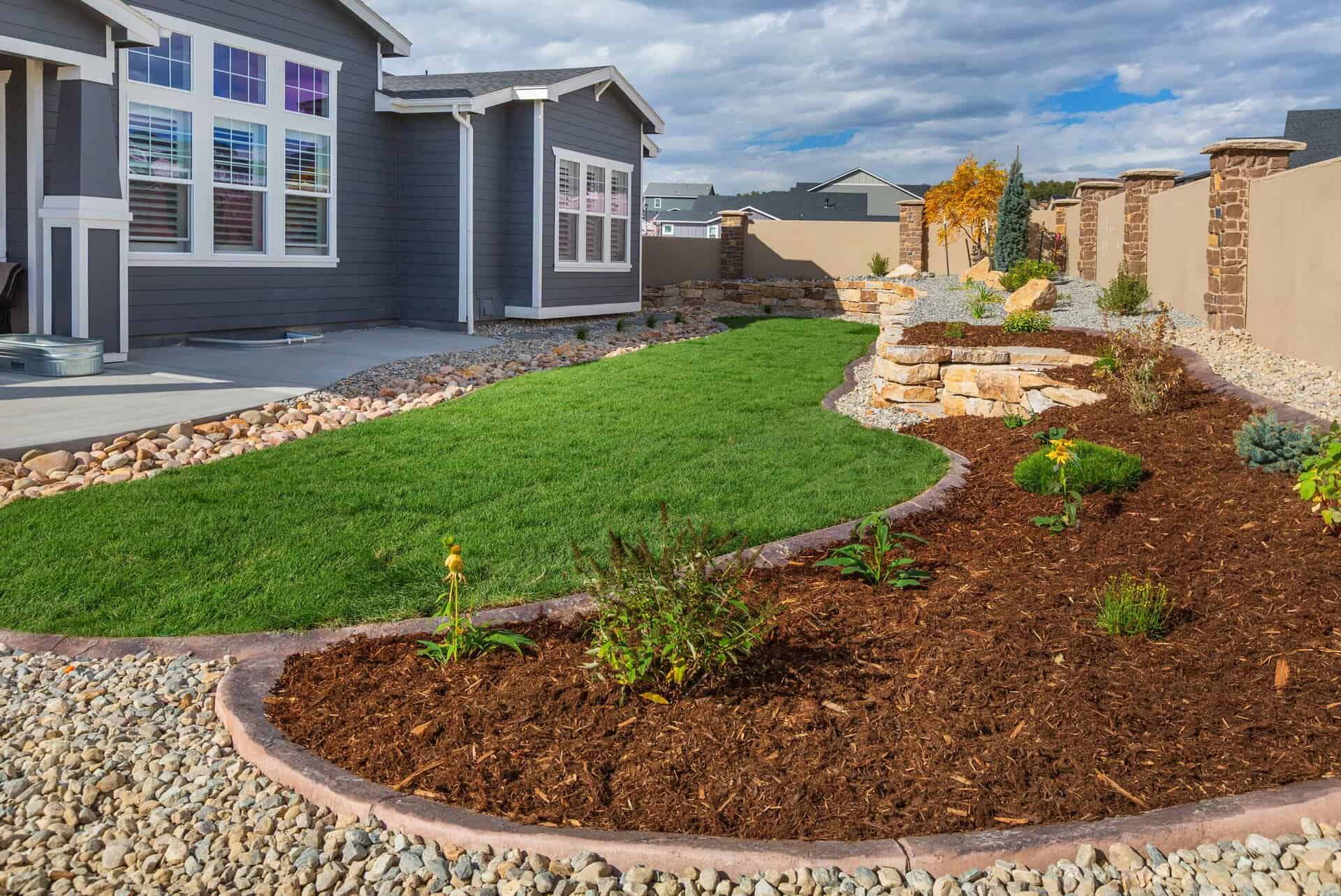 A landscaper-designed front yard features a curved green lawn, mulched garden beds, rock edging, and a modern gray house in the background.