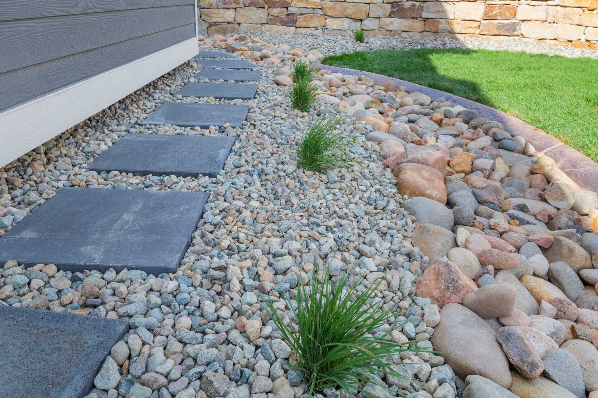 Close-up of a landscaped yard designed by a skilled landscaper, featuring square paver stones forming a path through gravel and small decorative plants, bordered by larger rocks and a curved grass edge.