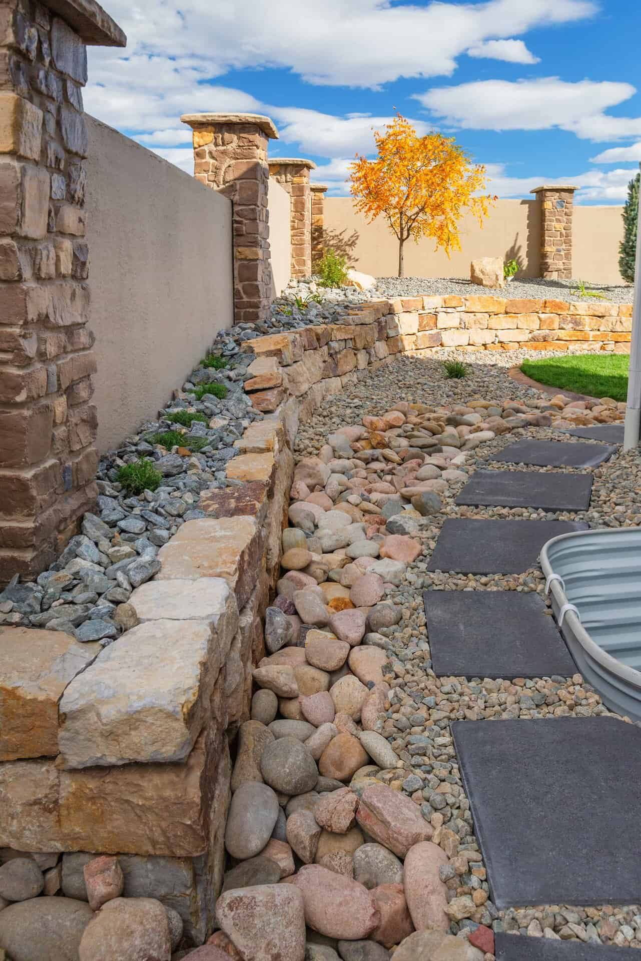 A landscaper-designed yard featuring stone retaining walls, a dry creek bed with large rocks, stepping stones, and a small tree with yellow leaves set against a blue sky.