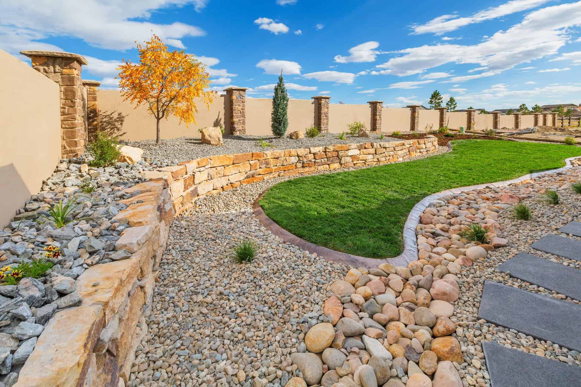 A landscaper-designed backyard with stone retaining walls, gravel beds, a small tree, lush green grass, and a stone pathway beneath a blue sky dotted with scattered clouds.