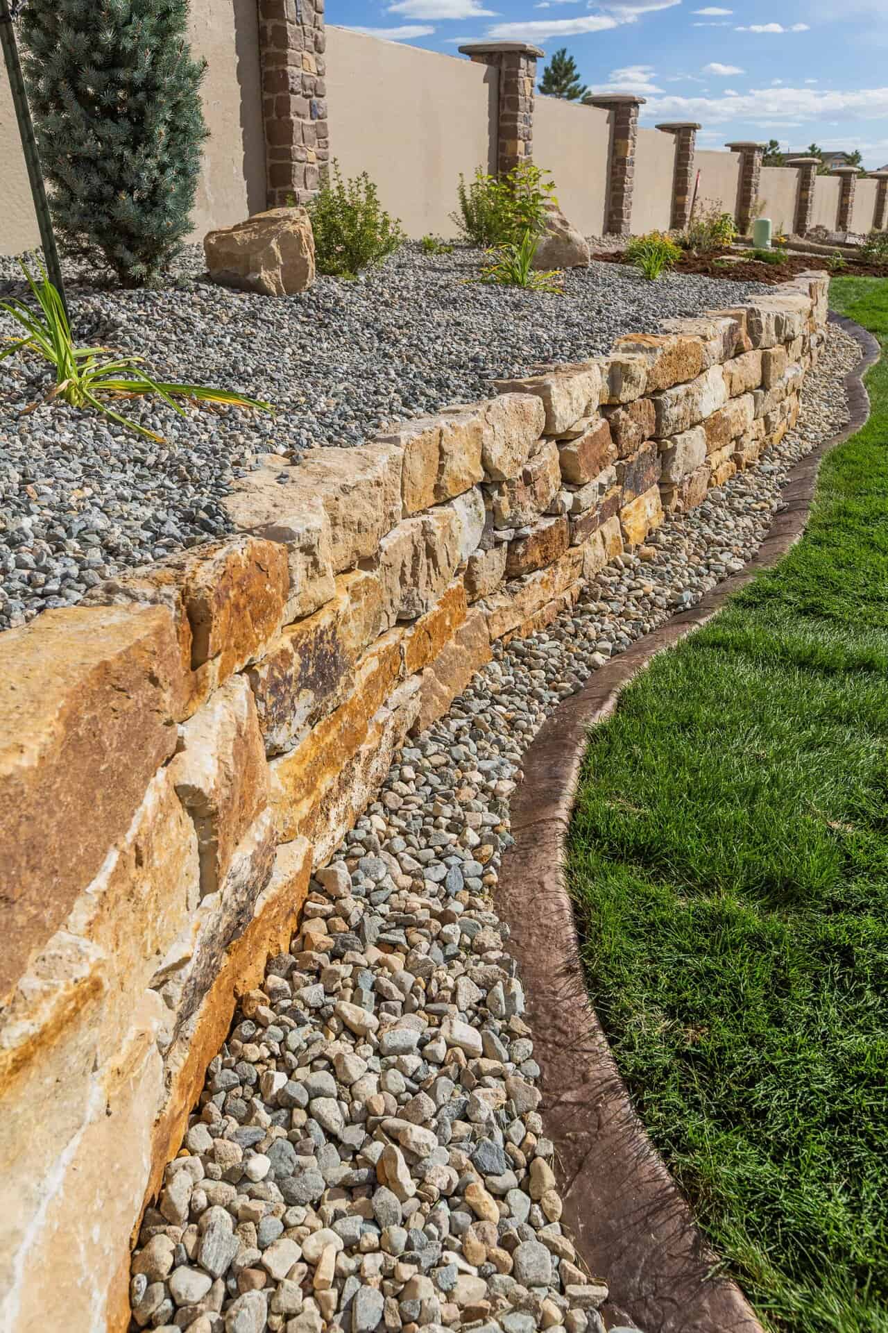 A stone retaining wall, crafted by a skilled landscaper, separates a gravel bed from a lush lawn, with small plants and a fence in the background under a blue sky.