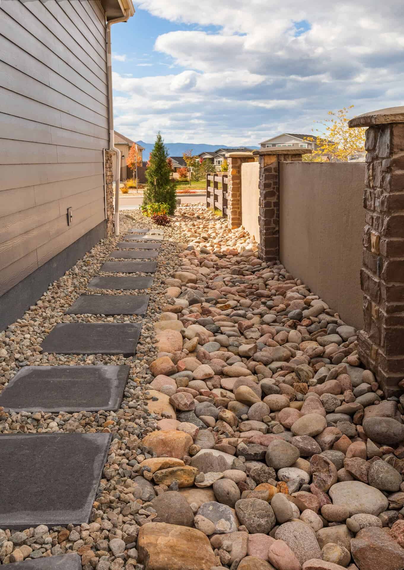 Side yard designed by a skilled landscaper, featuring a stone paver walkway and decorative river rocks, bordered by a house wall on one side and a stone fence on the other.