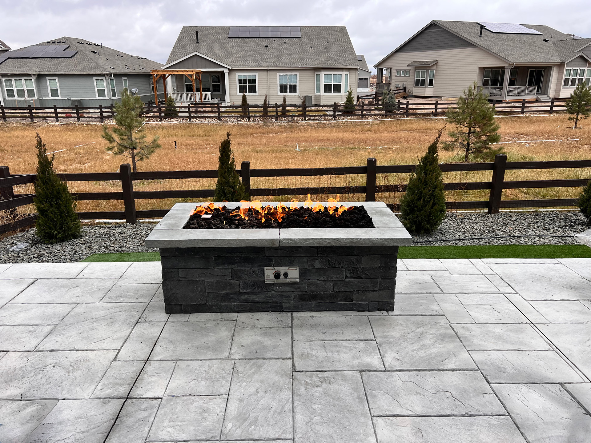 Rectangular outdoor gas fire pit with visible flames on a stone patio, surrounded by landscaped greenery, with houses and a wooden fence in the background.