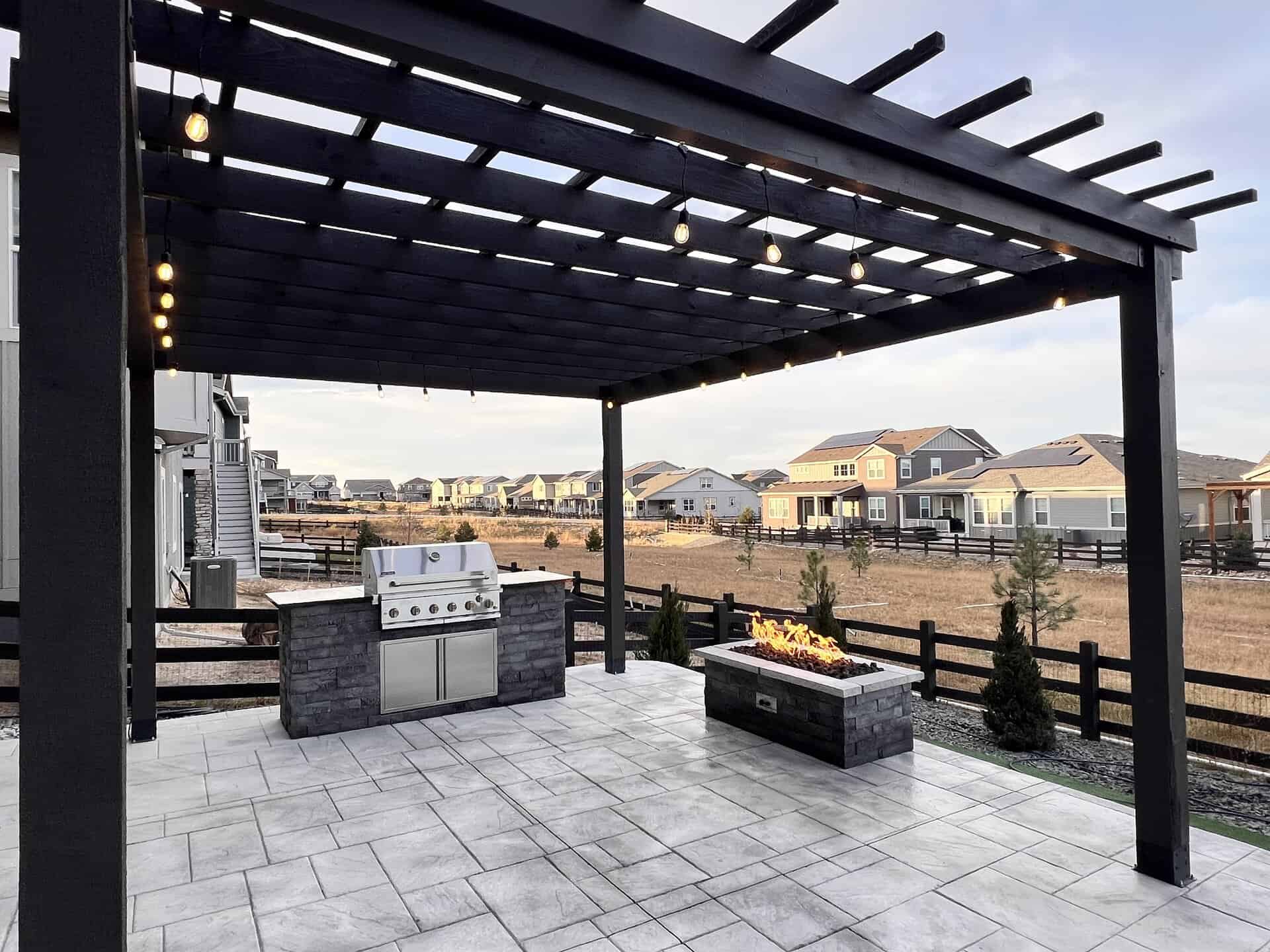 A modern outdoor patio with a pergola, built-in grill station, and rectangular fire pit; overlooking a suburban neighborhood and open field.