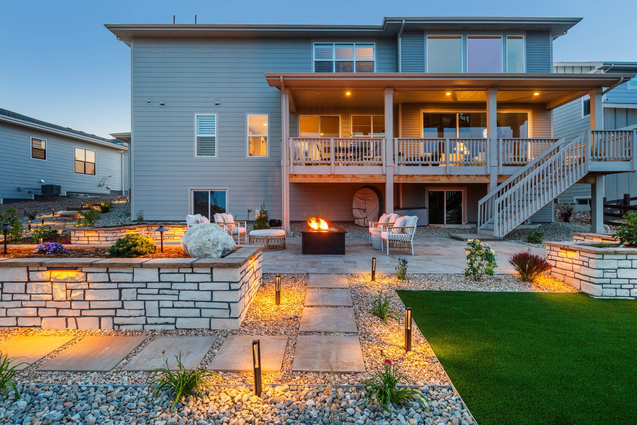 Modern backyard with stone pathways, illuminated landscaping, a central fire pit, outdoor seating, artificial turf, and a two-story house with a balcony and stairs in the background.